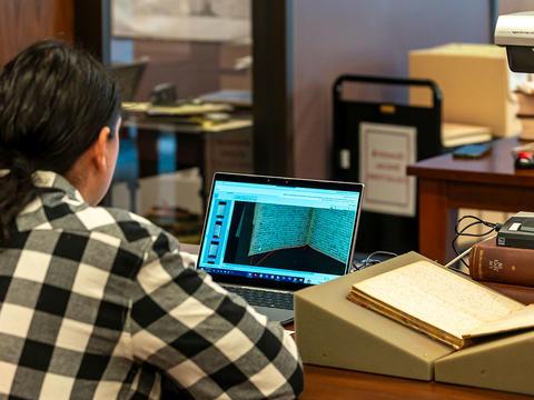 A man in a black and white checkered shirt uses an overhead scanner to process documents related to 贵格会教徒 run Native American Boarding Schools.